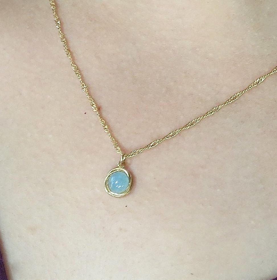 עשוי לכלול: שרשרת זהב עדינה הכוללת תליון קטן, אליפסה, עשוי אבן חן בצבע תכלת. לשרשרת שרשרת חבל עדינה ומפותלת. התליון משובץ היטב במסגרת זהב, מה שמדגיש את האלגנטיות העדינה שלו. שרשרת מינימליסטית זו מושלמת להוספת נגיעה של תחכום מאופק לכל תלבושת.