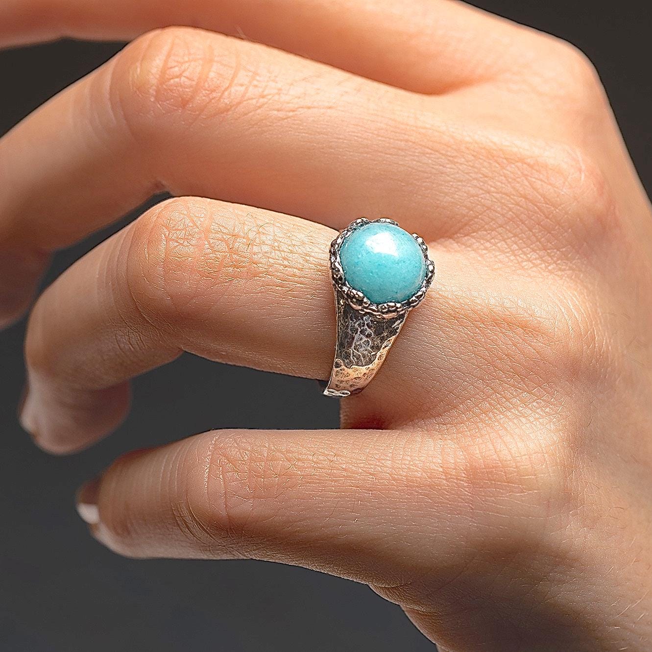 עשוי לכלול: טבעת כסף סטרלינג כוללת קבושון עגול משובץ במסגרת, עשוי אקוומרין טורקיז בהיר. קנה הטבעת בעל מרקם מרוקע. טבעת זו, שעוצבה בעבודת יד, מציגה עיצוב ייחודי ואבן חן איכותית.