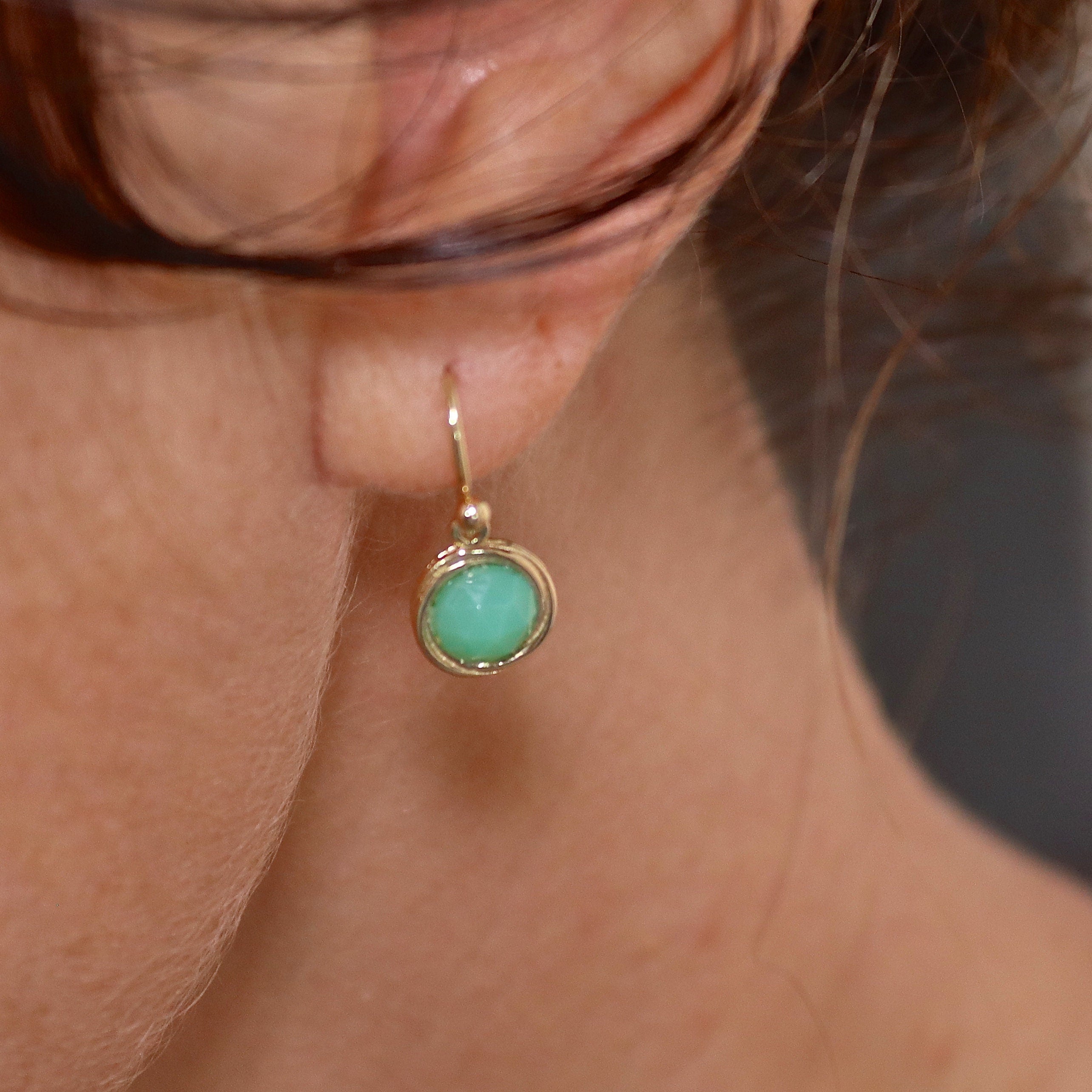 עשוי לכלול: תקריב של עגיל זהב בודד ובו אבן חן ירוקה בהירה, עגולה קטנה. לעגיל עיצוב עדין ופשוט, עם לולאת זהב קטנה בחלק העליון לתלייה. אבן החן חלקה ונראית כמו חיתוך קבושון. העגיל מוצג על רקע צוואר וכתף של אדם, עם מעט שיער חום כהה גלוי.