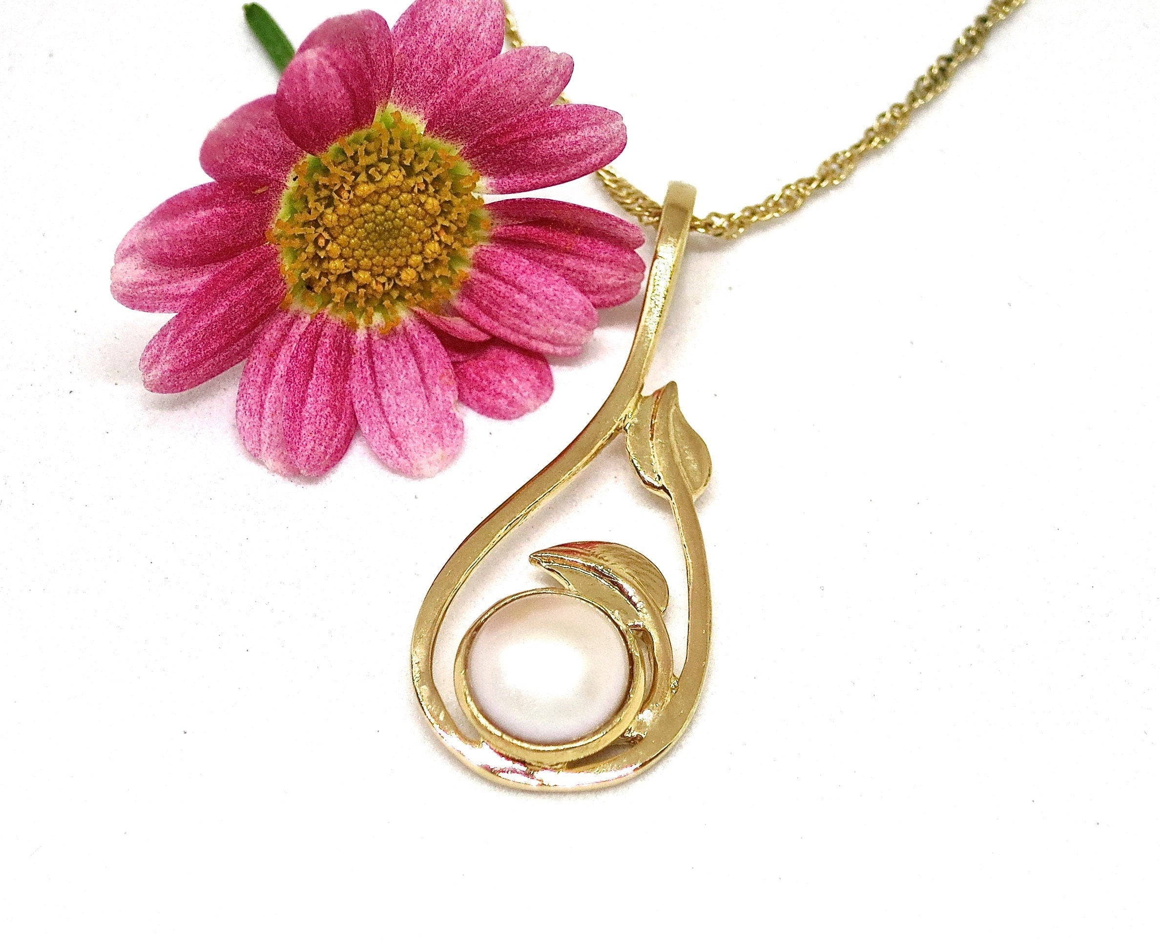 עשוי לכלול: שרשרת תליון זהב בעלת עיצוב בצורת אגס עם נגיעות עדינות של עלים. פנינה עגולה אחת בצבע לבן-שבור משובצת במסגרת המעוטרת של התליון. השרשרת מוצגת על רקע לבן, מעוטרת בחינניות ורודות עזות.