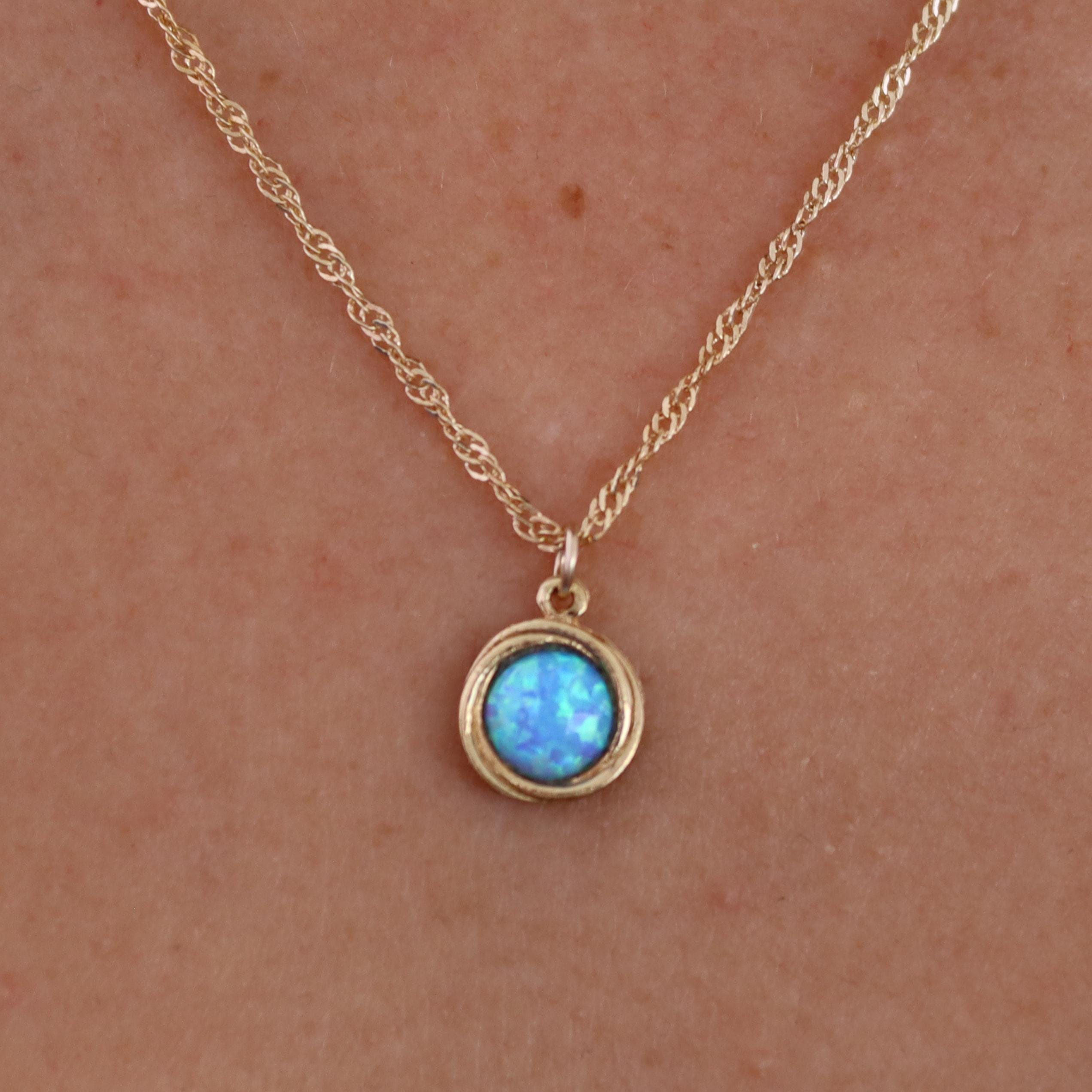 עשוי לכלול: שרשרת חבל זהב עדינה הכוללת תליון עגול קטן. התליון מציג אבן חן אופל תכלת, מוקפת במסגרת זהב. האופל מציג משחק צבעים עדין, האופייני לאבן חן זו. השרשרת פשוטה ואלגנטית, מתאימה לענידה יומיומית או לאירועים מיוחדים. העיצוב הכללי מינימליסטי ומעודן.