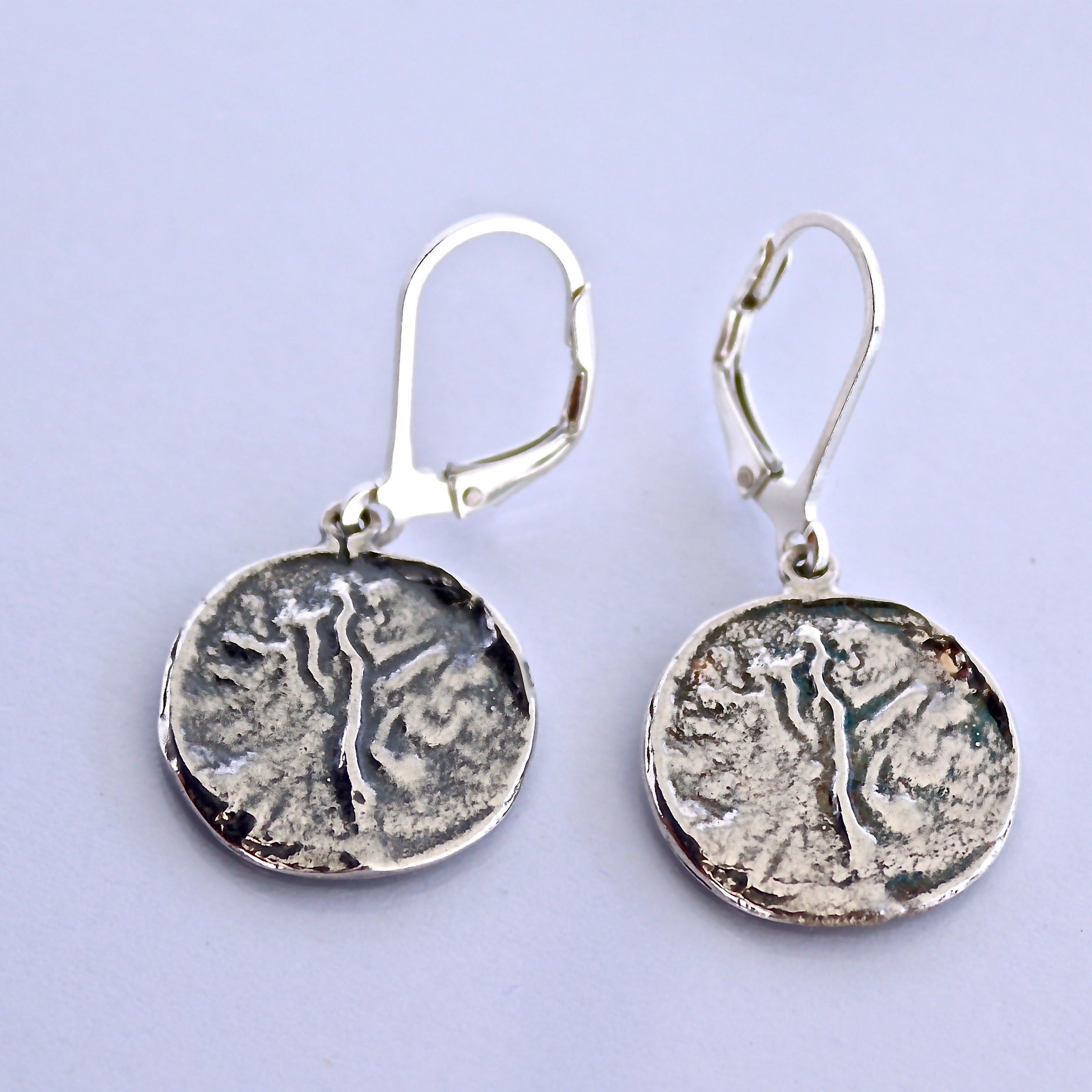 עשויים לכלול: זוג עגילי כסף בעיצוב עגול הכולל צללית עץ בעלת מרקם. לעגילים יש סוגר קלאפס (Leverback) מנגנון סגירה.