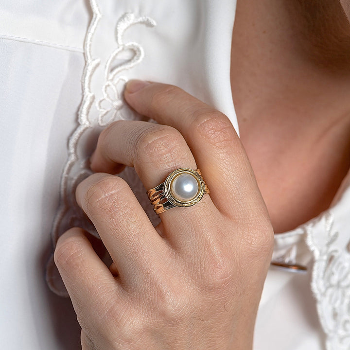 עשוי לכלול: טבעת זהב עם פנינה לבנה גדולה משובצת במסגרת עגולה. לטבעת רצועה בעלת טקסטורה עם שלושה פסים בולטים.