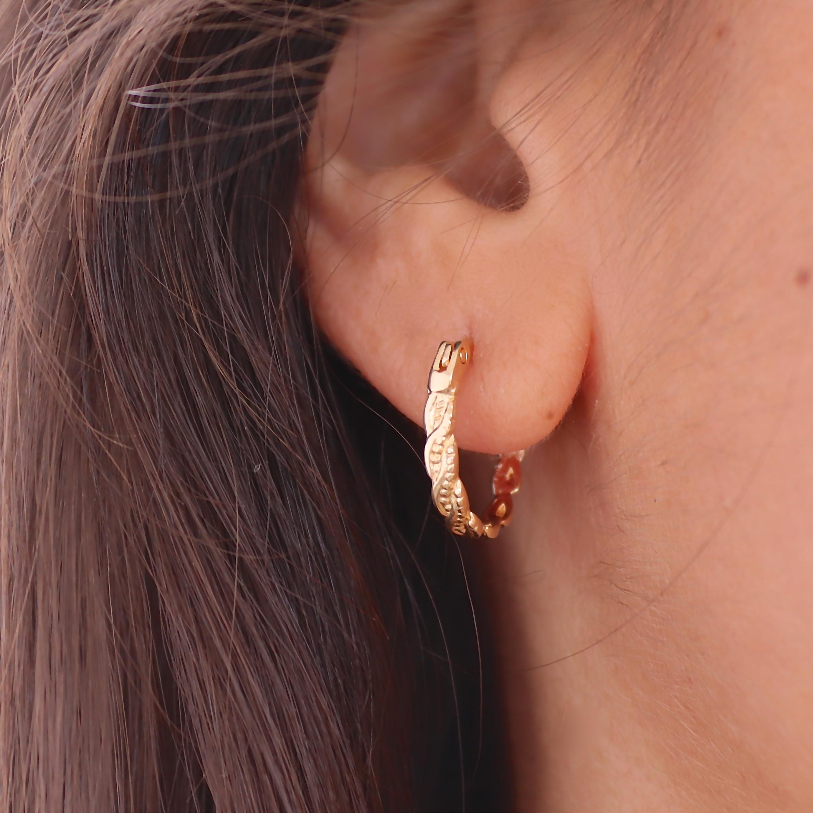 עשוי לכלול: עגיל חישוק זהב עם עיצוב טקסטורה. העגיל הוא בצורת עגולה קטנה וכולל דוגמה של צורות חוזרות דמויות עלים. העגיל נלבש על האוזן, כאשר העיצוב נראה לעין. העגיל עשוי מתכת מבריקה בצבע זהב.