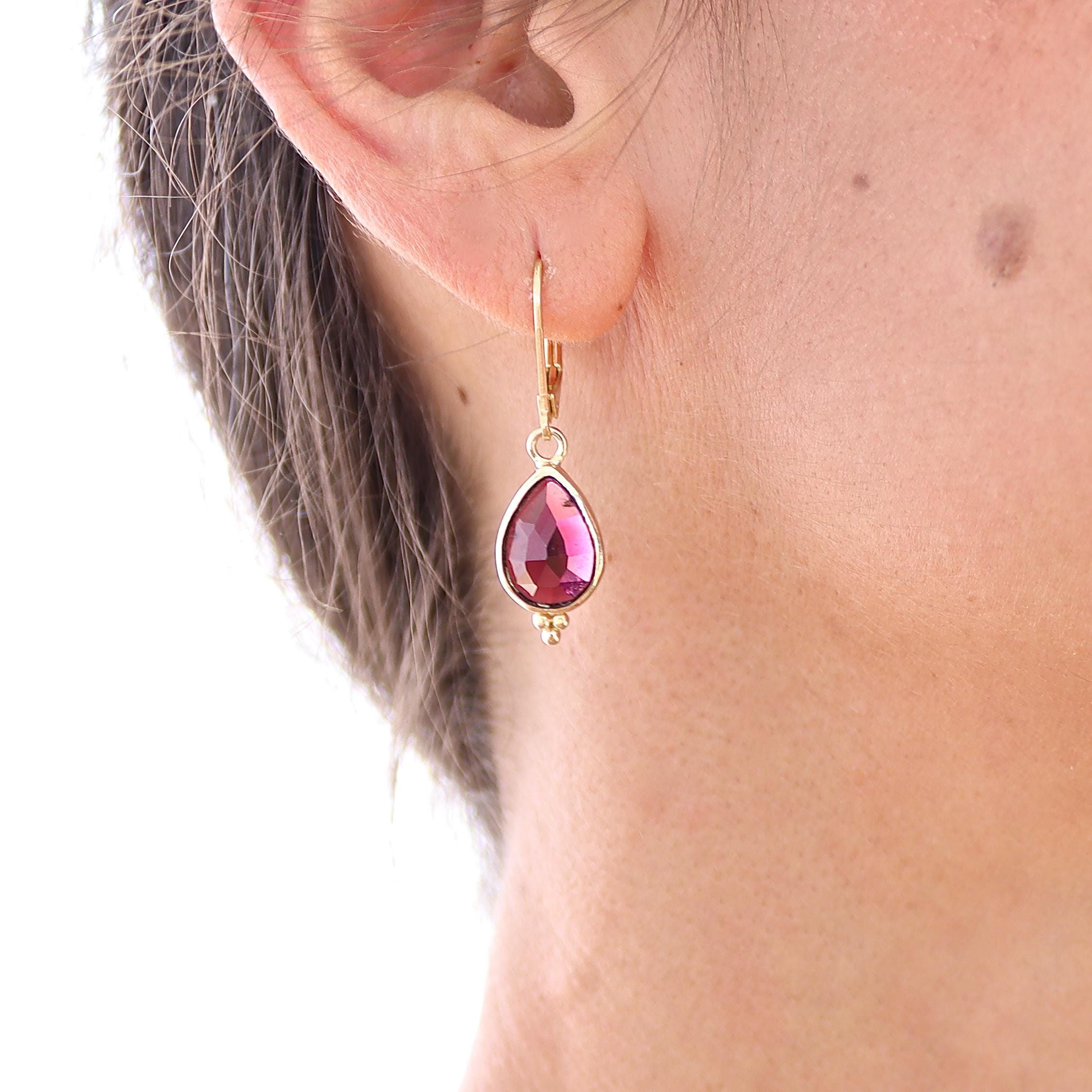 עשוי לכלול: עגיל בצבע זהב עם אבן חן ורודה בצורת דמעה. אבן החן משובצת במסגרת זהב ובעלת משטח פאותי. העגיל תלוי על אבזם מנוף אחורי. בתחתית אבן החן, ישנם שלושה חרוזי זהב קטנים.