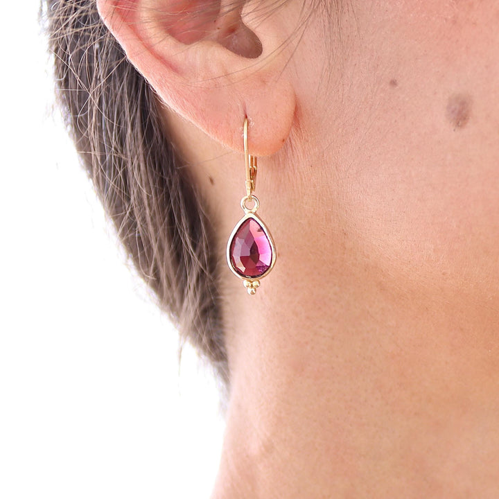 עשוי לכלול: עגיל בצבע זהב עם אבן חן ורודה בצורת דמעה. אבן החן משובצת במסגרת זהב ובעלת משטח פאותי. העגיל תלוי על אבזם מנוף אחורי. בתחתית אבן החן, ישנם שלושה חרוזי זהב קטנים.