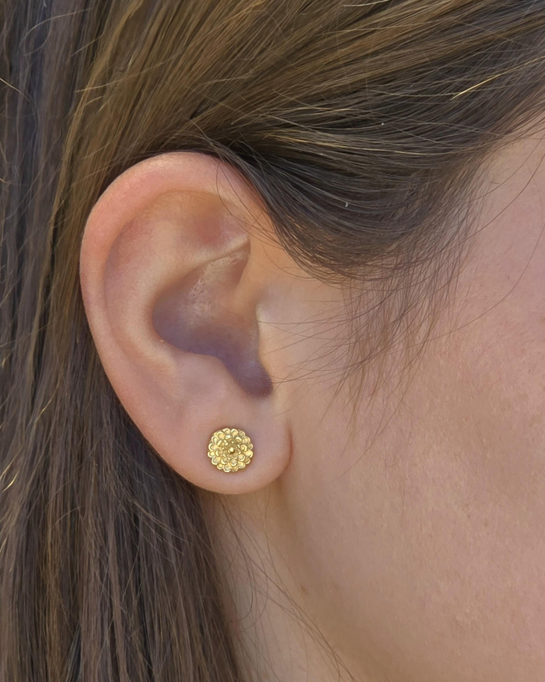 עשוי לכלול: תקריב של עגיל זהב צמוד על אוזן. העגיל עגול עם עיצוב פרחוני. העגיל קטן ויושב ישירות על תנוך האוזן. העור סביב האוזן בגוון בהיר והשיער חום.