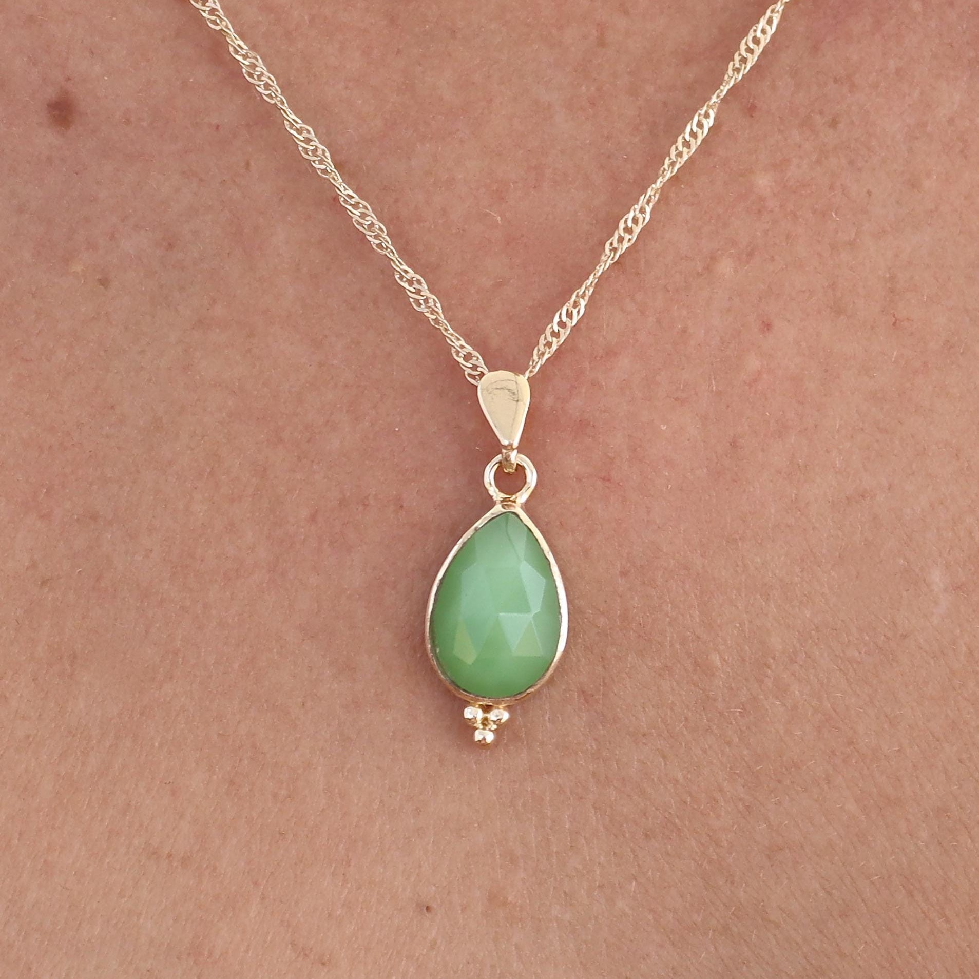 עשוי לכלול: שרשרת זהב עדינה הכוללת תליון אבן חן ירוקה בהירה בצורת אגס. התליון משובץ בעדינות בחרוזי זהב קטנים בתחתיתו. שרשרת הזהב הדקה של השרשרת מוסיפה לפשטותה האלגנטית, מה שהופך אותה לאביזר מושלם לכל אירוע. הגוון הירוק הבהיר והרך של אבן החן משלים את הזהב בצורה יפהפייה, ויוצר מראה מתוחכם ומאופק. העיצוב הכללי מינימליסטי ושיקי, מתאים ללבוש יומיומי או לאירועים מיוחדים.