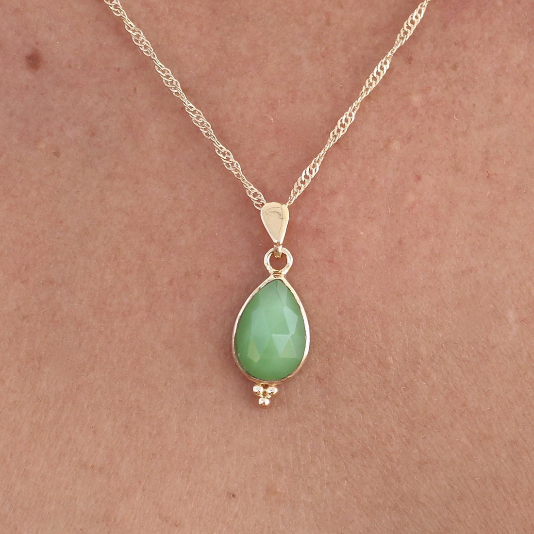 עשוי לכלול: שרשרת זהב עדינה הכוללת תליון אבן חן ירוקה בהירה בצורת אגס. התליון משובץ בעדינות בחרוזי זהב קטנים בתחתיתו. שרשרת הזהב הדקה של השרשרת מוסיפה לפשטותה האלגנטית, מה שהופך אותה לאביזר מושלם לכל אירוע. הגוון הירוק הבהיר והרך של אבן החן משלים את הזהב בצורה יפהפייה, ויוצר מראה מתוחכם ומאופק. העיצוב הכללי מינימליסטי ושיקי, מתאים ללבוש יומיומי או לאירועים מיוחדים.