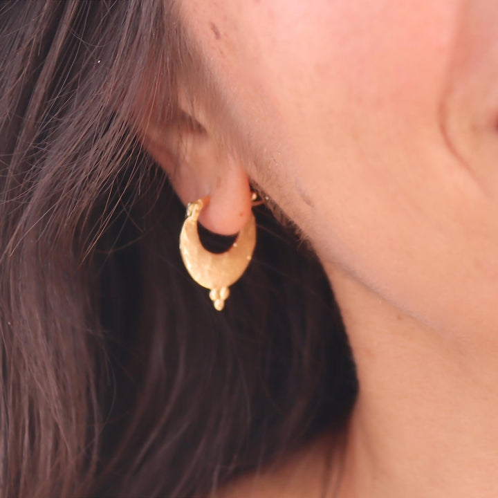 עשוי לכלול: עגיל בצבע זהב בצורת סהר. לעגיל משטח מחוספס ופרט קטן ומתנדנד בתחתית. העגיל נלבש על אוזנו של אדם, כאשר דגש הוא על עיצוב ופרטי התכשיט.