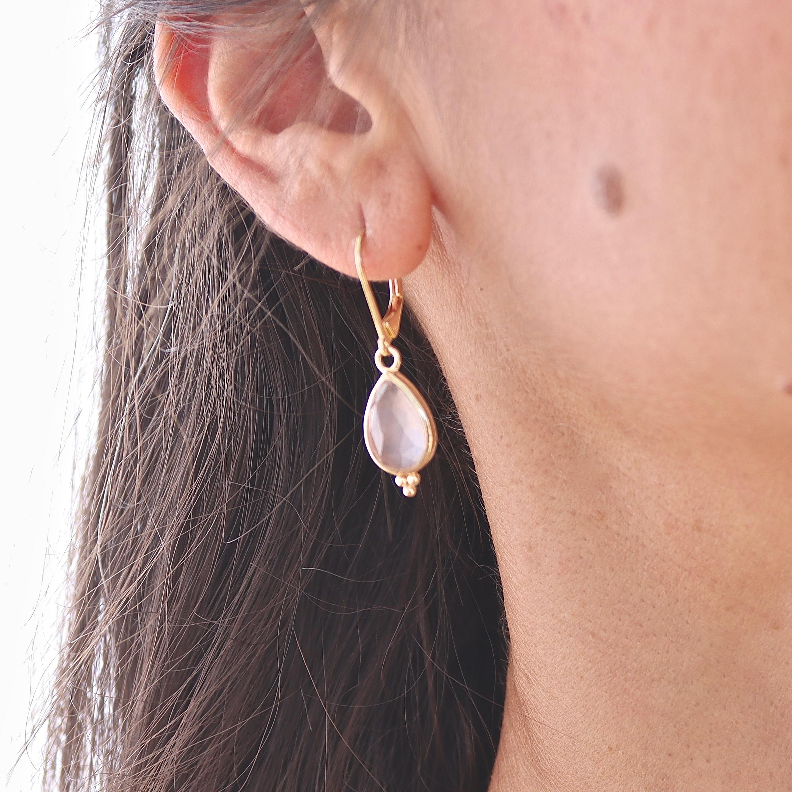 עשוי לכלול: עגיל בצבע זהב עם אבן חן ורודה בהירה בצורת אגס. אבן החן תלויה על וו קטן ומעוקל. שלושה חרוזי זהב קטנים נמצאים בתחתית אבן החן. העגיל נלבש על אוזן אדם.