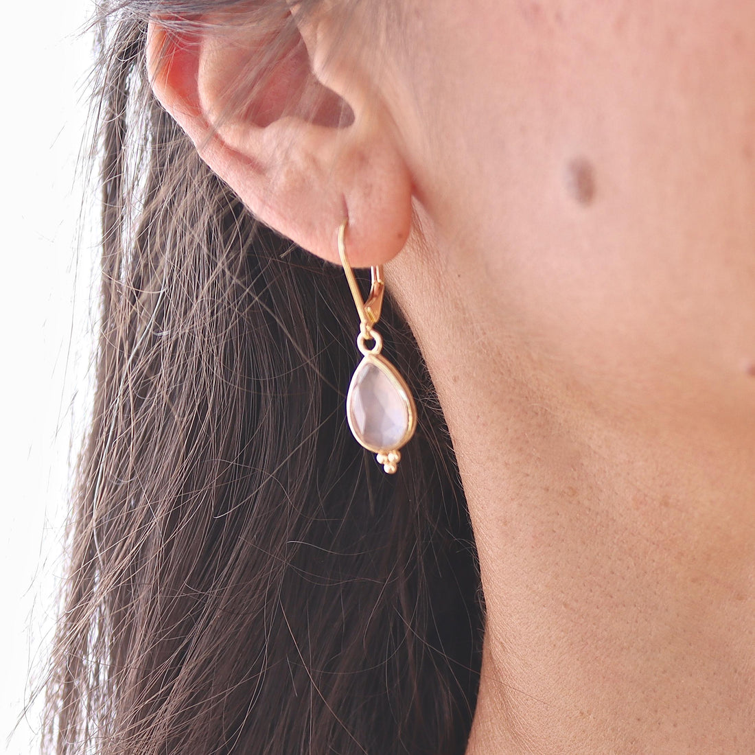 עשוי לכלול: עגיל בצבע זהב עם אבן חן ורודה בהירה בצורת אגס. אבן החן תלויה על וו קטן ומעוקל. שלושה חרוזי זהב קטנים נמצאים בתחתית אבן החן. העגיל נלבש על אוזן אדם.