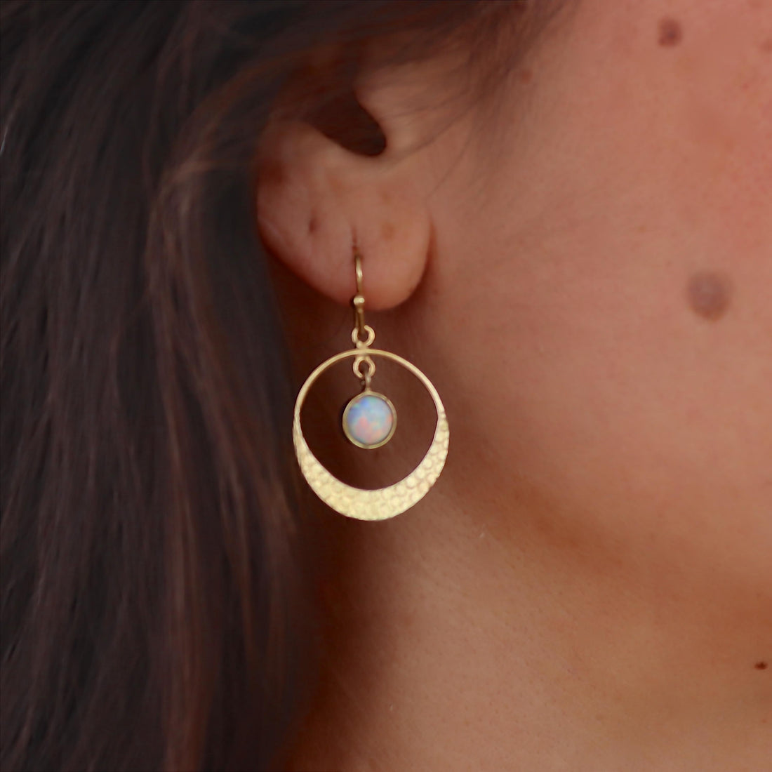 עשוי לכלול: עגיל בצבע זהב עם עיצוב עגול. העגיל כולל טבעת חיצונית בעלת טקסטורה ועיגול פנימי קטן יותר המחזיק אבן לבנה ועגולה. העגיל תלוי על וו קטן (סוגר וו), המציג סגנון מינימליסטי ואלגנטי.