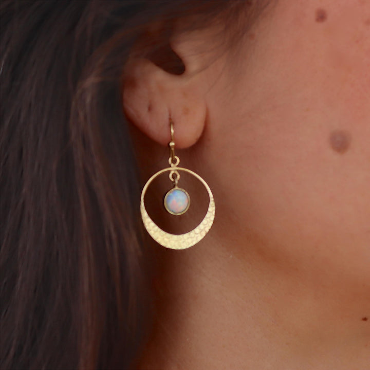 עשוי לכלול: עגיל בצבע זהב עם עיצוב עגול. העגיל כולל טבעת חיצונית בעלת טקסטורה ועיגול פנימי קטן יותר המחזיק אבן לבנה ועגולה. העגיל תלוי על וו קטן (סוגר וו), המציג סגנון מינימליסטי ואלגנטי.