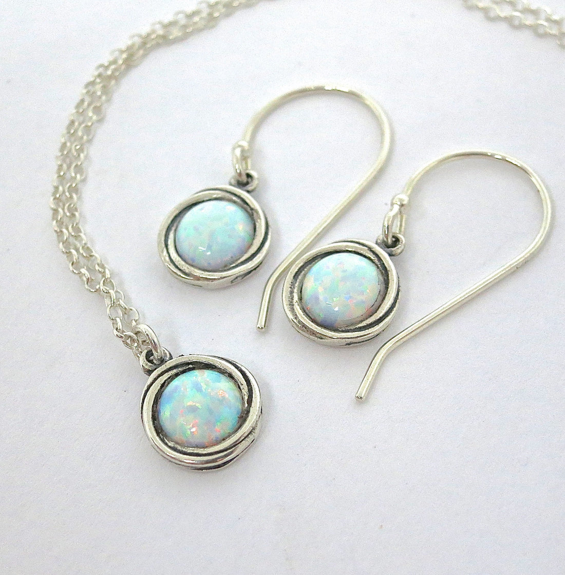 עשוי לכלול: סט שרשרת ועגילים מכסף הכולל תליוני אופל. לשרשרת שרשרת עדינה עם תליון עגול. העגילים הם בצורת טיפה עם וו מעוגל (סוגר וו). כל פריט מציג אבן אופל עגולה וססגונית משובצת במסגרת כסף מעוותת.