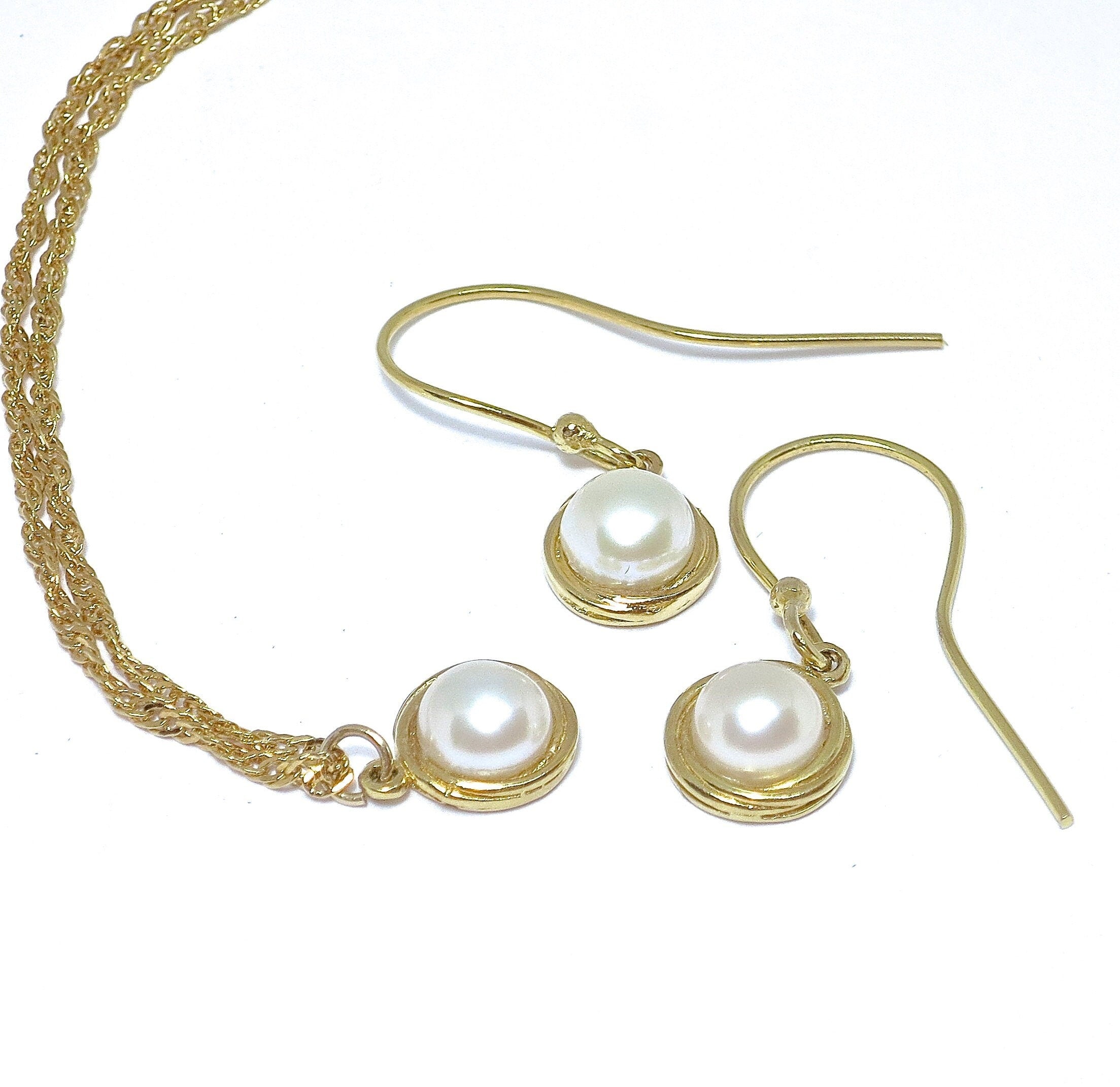 עשוי לכלול: סט שרשרת ועגילים מזהב הכולל פנינים קטנות, עגולות, בצבע לבן-שבור. השרשרת עשויה משרשרת חבל כפולה ועדינה. כל עגיל ותליון משובצים בזהב פשוט. הפנינים נראות חלקות ומבריקות. סט התכשיטים אלגנטי ומינימליסטי בסגנון.