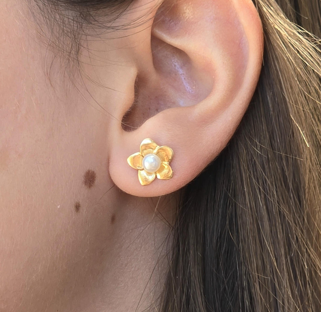 עשוי לכלול: עגיל פרחים בצבע זהב עם מרכז פנינה. העגיל בצורת פרח עם חמישה עלי כותרת. הפנינה היא חרוז לבן, עגול, קטן. העגיל נמצא על אוזן של אדם.