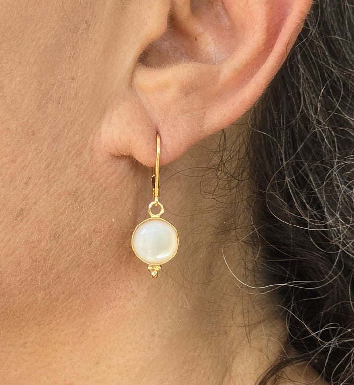 עשוי לכלול: עגיל בצבע זהב עם אבן עגולה, לבנה, דמוית פנינה. לעגיל יש מנוף אחורי מגנטי ומקבץ קטן ודקורטיבי של שלושה חרוזי זהב בתחתית האבן. העגיל מוצג על אוזן אדם.