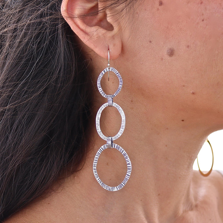 עשוי לכלול: עגילי כסף תלויים עם שלושה עיגולים פתוחים בעלי טקסטורה. העיגולים מחוברים בקו אנכי, כאשר העיגול העליון מחובר לוו (וו) לאוזן. לעגילים מראה מחורר או חרוט, היוצר משטח בעל טקסטורה. העיגולים בצבע כסף בוהק.