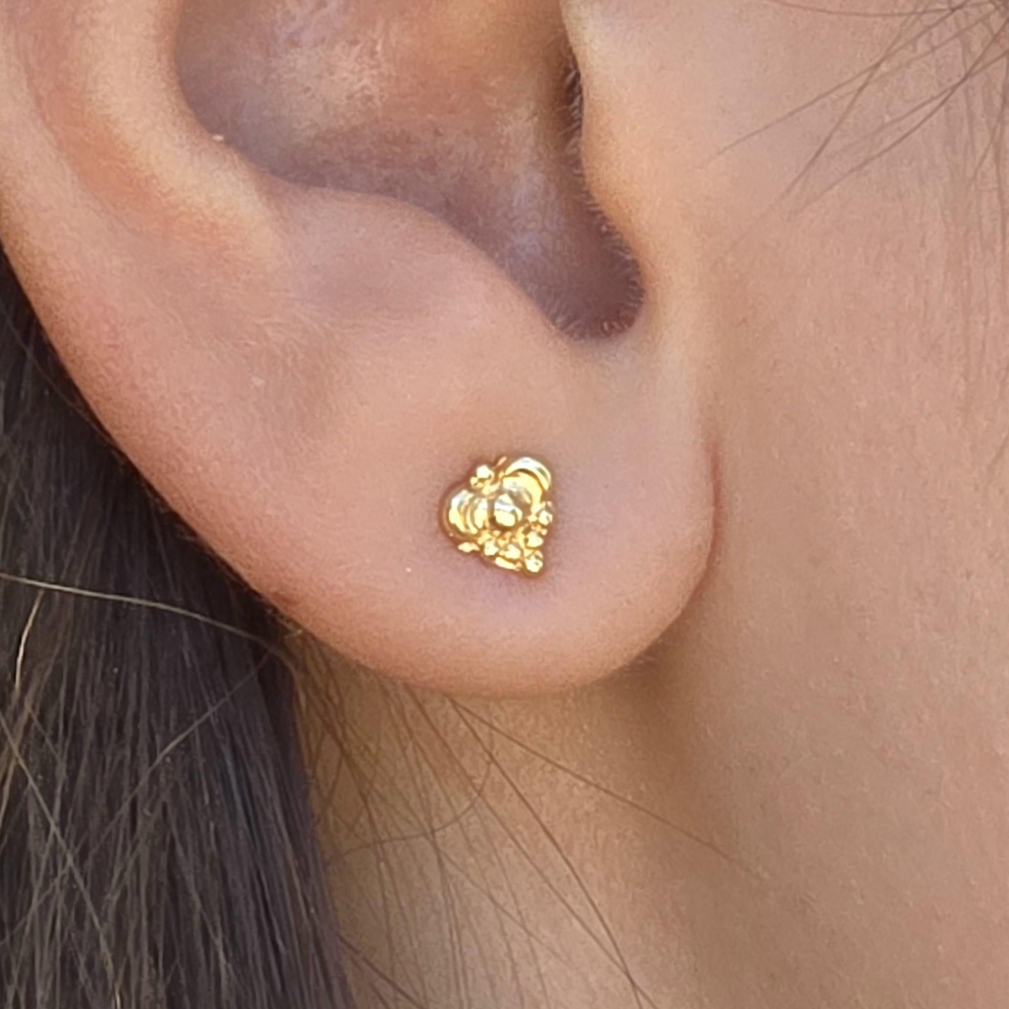 עשוי לכלול: עגיל קטן בצבע זהב נראה על אוזן. לעגיל עיצוב אשכול, עם צורה מעוגלת ופרטים מורכבים. העגיל נראה עשוי מתכת וממוקם על תנוך האוזן. גוון העור בהיר.