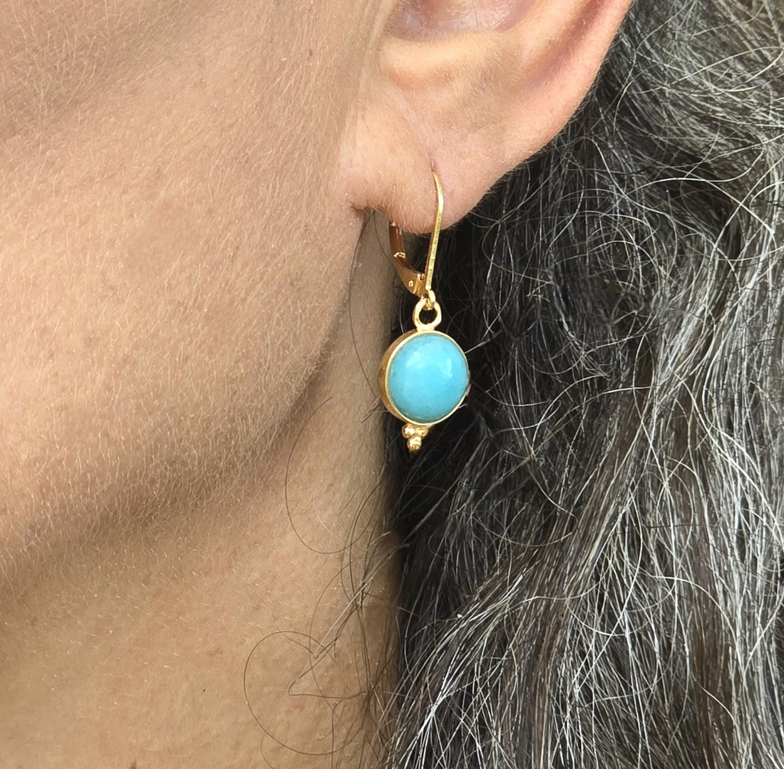 עשוי לכלול: עגיל בצבע זהב עם אבן עגולה בצבע תכלת. לעגיל גב מנוף "מנגנון סגירה" ועיצוב דקורטיבי קטן בתחתית. העגיל תלוי על אוזן.