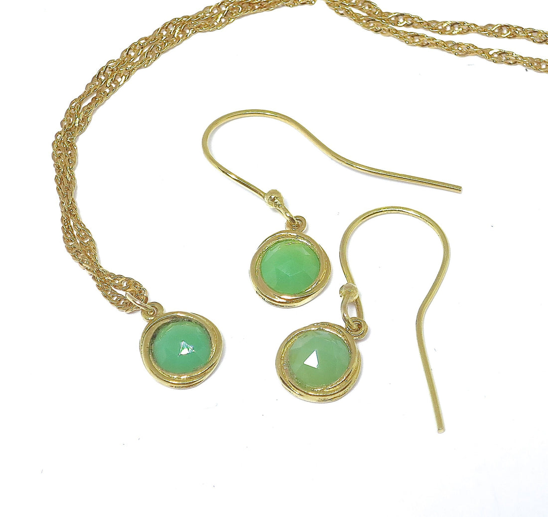 עשוי לכלול: שרשרת ועגילים מזהב משובצים באבני חן עגולות בצבע ירוק בהיר. לשרשרת שרשרת עדינה ומפותלת, והעגילים הם בסגנון טיפה פשוט. האבנים נראות בחיתוך קבושון ובעלות איכות עדינה ושקופה. התכשיטים אלגנטיים ומינימליסטיים, מתאימים לענידה יומיומית או לאירועים מיוחדים. הזהב הוא בגוון צהוב חם.