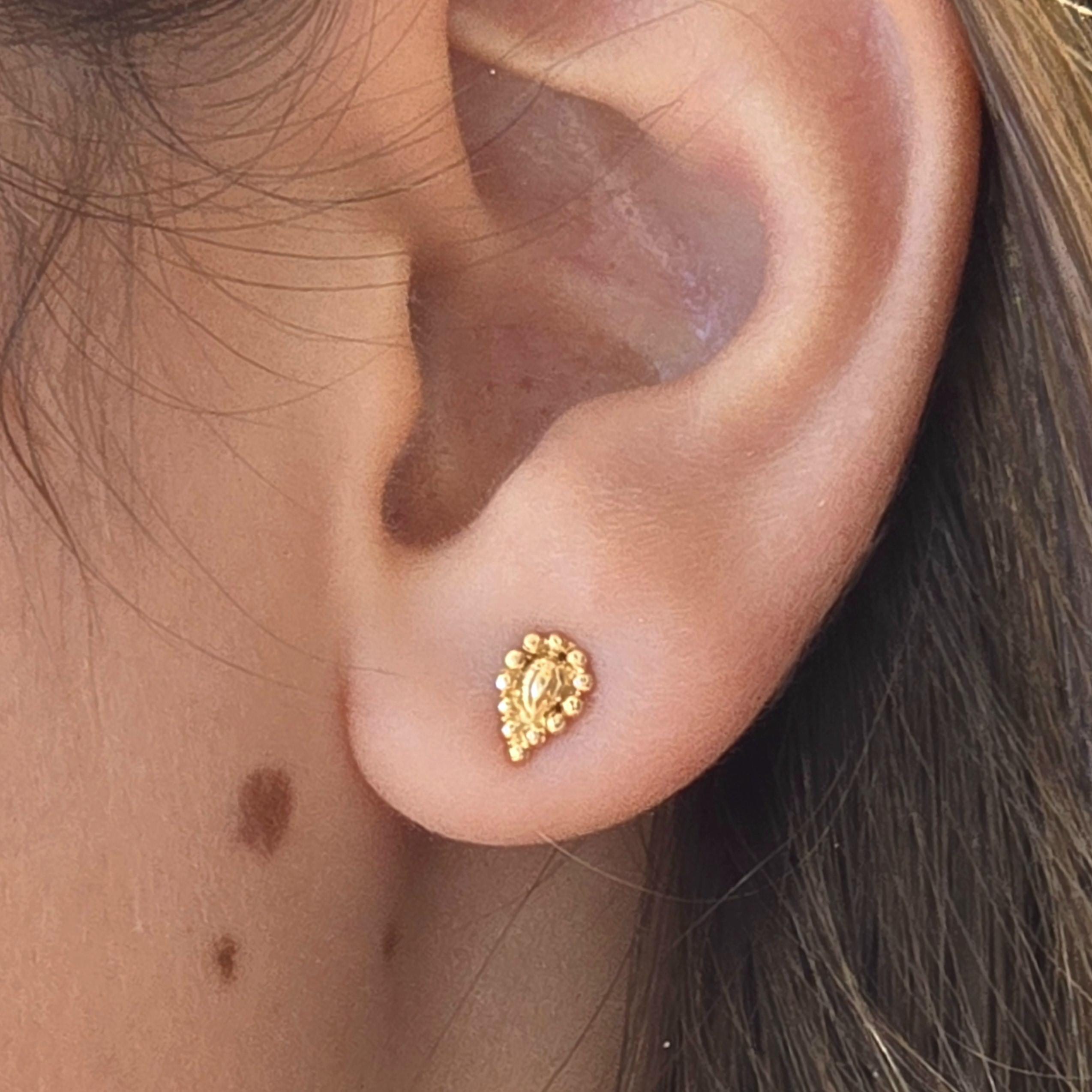 עשוי לכלול: עגיל קטן בצבע זהב בצורת דמעה. העגיל משובץ בחרוזים עגולים קטנים, היוצרים משטח מחוספס. העגיל ממוקם על אוזן, ומציג את גודלו ועיצובו. לעגיל מראה מבריק.