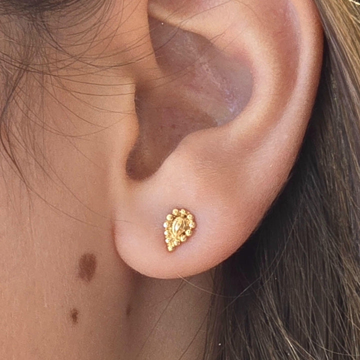 עשוי לכלול: עגיל קטן בצבע זהב בצורת דמעה. העגיל משובץ בחרוזים עגולים קטנים, היוצרים משטח מחוספס. העגיל ממוקם על אוזן, ומציג את גודלו ועיצובו. לעגיל מראה מבריק.