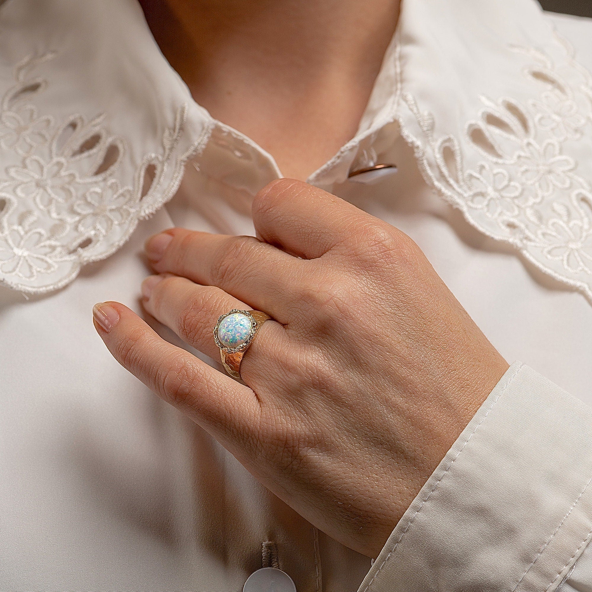 עשוי לכלול: טבעת זהב כוללת אבן חן אופל עגולה בצבע תכלת. פני השטח של האופל מציגים נצנוץ ומרקם עדינים. טבעת הטבעת עשויה מזהב, ומציגה גימור מעט מרוקע או בעל מרקם. המסגרת סביב האופל מפורטת עם נגיעות קטנות ונוצצות.