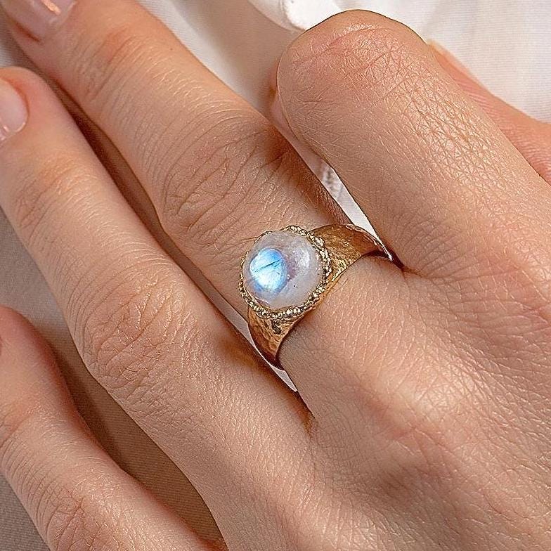 עשוי לכלול: טבעת זהב עם אבן חן גדולה, עגולה ולבנה. לאבן החן ברק כחול ססגוני.