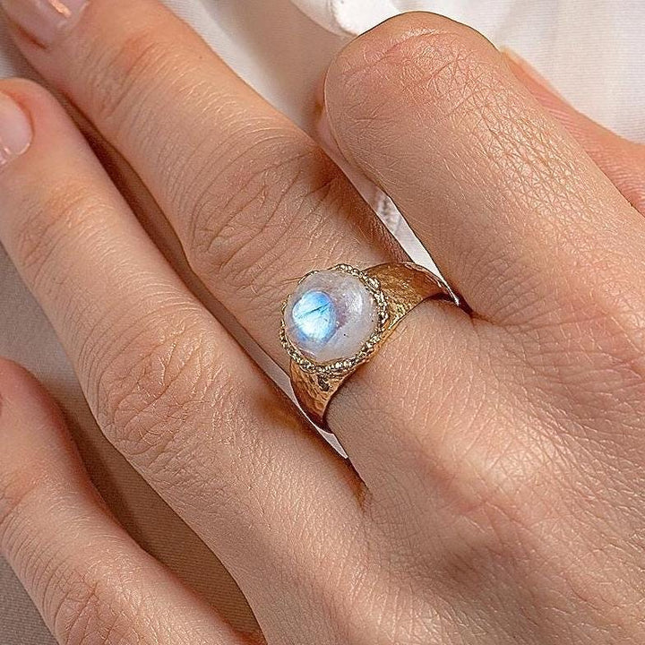 עשוי לכלול: טבעת זהב עם אבן חן גדולה, עגולה ולבנה. לאבן החן ברק כחול ססגוני.